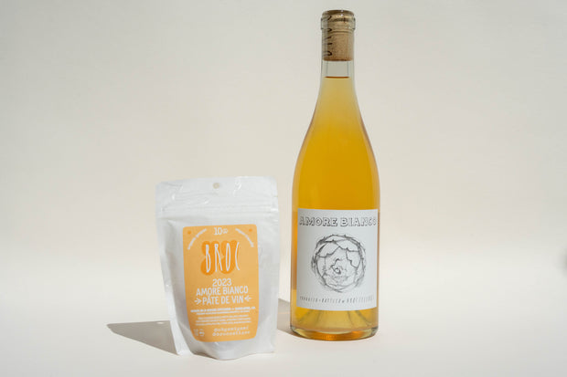 A bottle of Broc Cellars Amore Bianco + Yami Yami Pâte de Vin stands beside a clear pouch labeled “2023 Amore Bianco Pâté de Vin” against a light background.