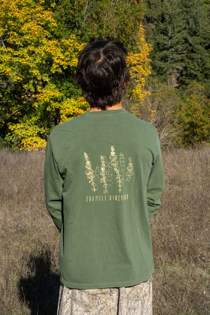 A person in a field faces away, wearing the Fox Hill Organic Long Sleeve Shirt by Broc Cellars—a green unisex cotton top with botanical illustrations and “Fox Hill Vineyard” on the back. Trees with yellow leaves, designed by Marta Elise Johansen, frame the scene.