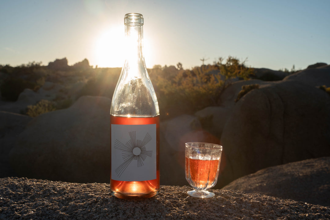 A clear bottle of Broc Cellars 2024 O’Sunshine rosé and a filled wine glass rest on a rock at sunset, surrounded by desert plants and boulders, as the setting sun hints at the wine’s bright cherry notes.