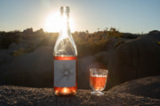 A clear bottle of Broc Cellars 2024 O’Sunshine rosé and a filled wine glass rest on a rock at sunset, surrounded by desert plants and boulders, as the setting sun hints at the wine’s bright cherry notes.