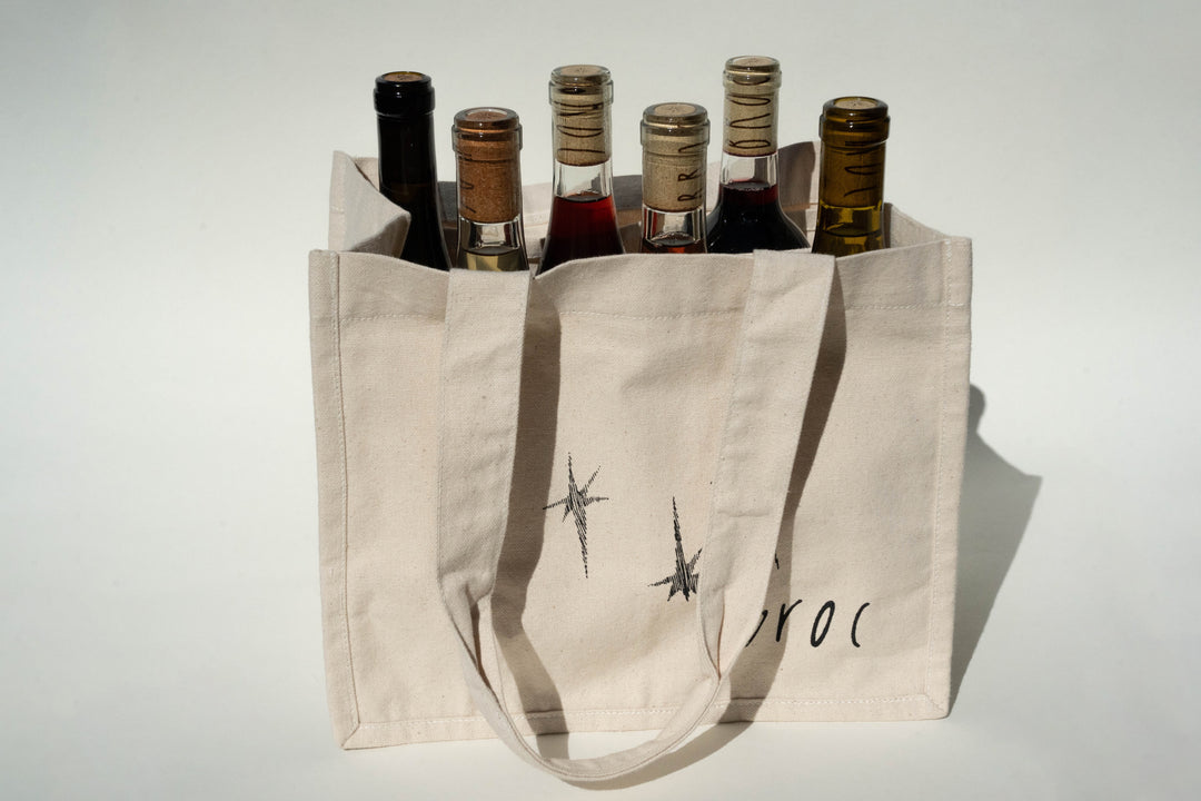 The Broc Cellars Broc Bottle Tote is a beige canvas tote with two handles and compartments for six wine bottles. Made in America, it features black star designs and partial text on the front against a plain, well-lit background.