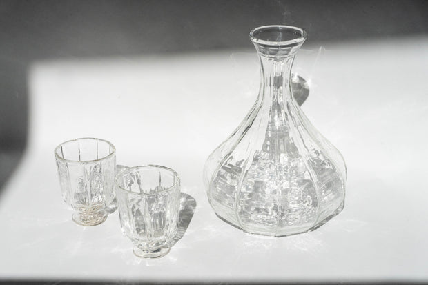 A clear Broc Cellars Decanter + Glass Set with a textured surface stands beside two matching glass cups on a white surface, illuminated to create mesmerizing reflections and shadows.