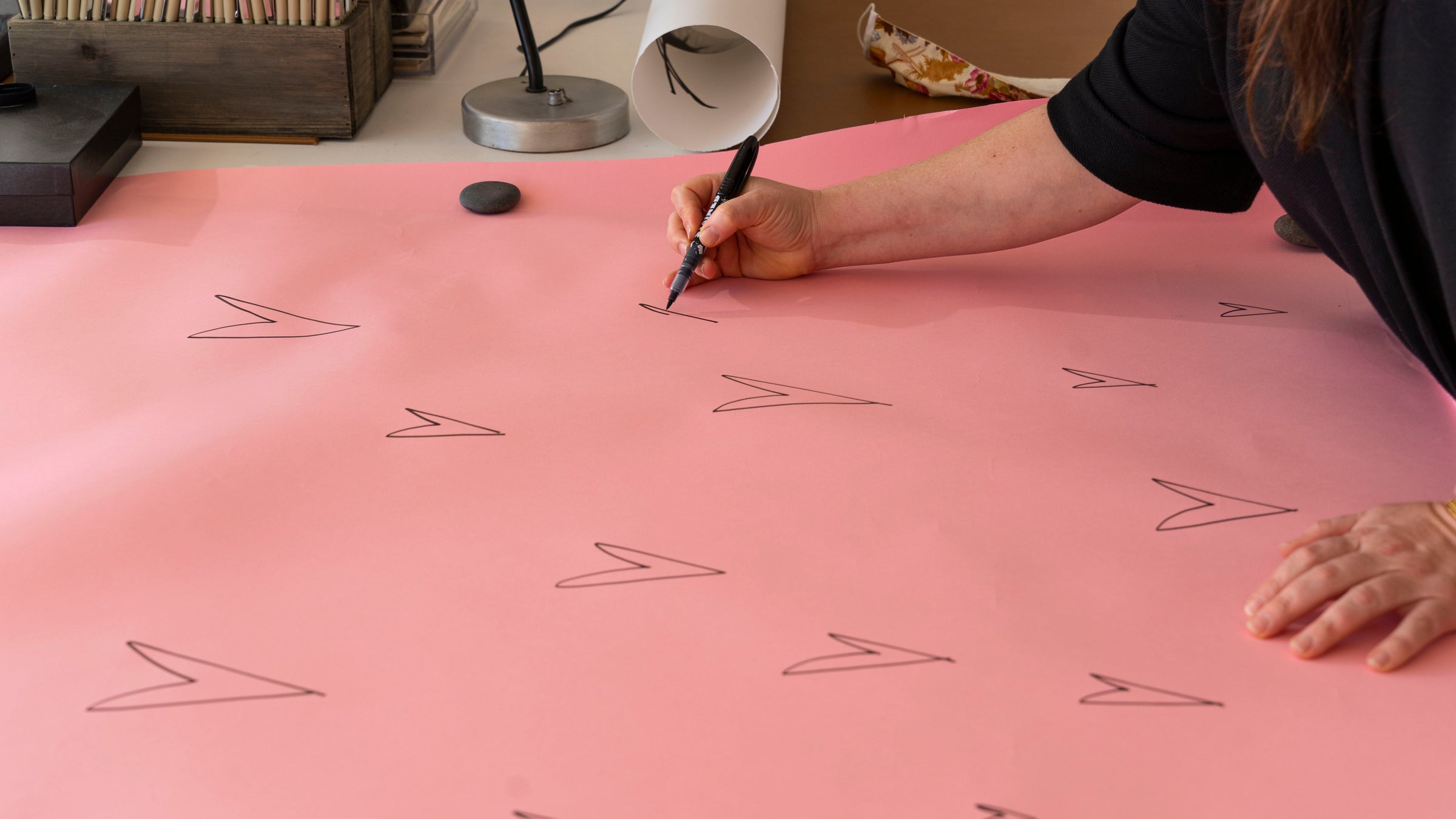 A person draws multiple bird footprints with a black marker on a large sheet of pink paper, with stones and objects on the table nearby.