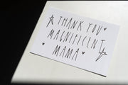 A Broc Cellars Mothers Day Packs card features handwritten THANK YOU MAGNIFICENT MAMA in uppercase letters, decorated with small hearts and stars, and placed on a light-colored surface.
