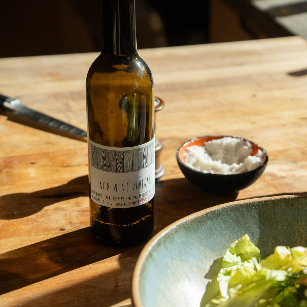 A bottle of Broc Cellars Camino Red Wine Vinegar rests on a wooden table beside a small bowl of coarse salt and a ceramic bowl of lettuce leaves, all illuminated by warm sunlight.