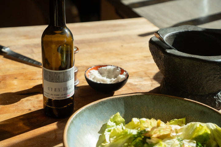 A bottle of CAMINO RED WINE VINEGAR from Broc Cellars sits on a wooden table alongside a small bowl of salt and a green salad in a ceramic bowl. A stone mortar with pestle is partially visible, and a knife rests in the background, all bathed in sunlight.