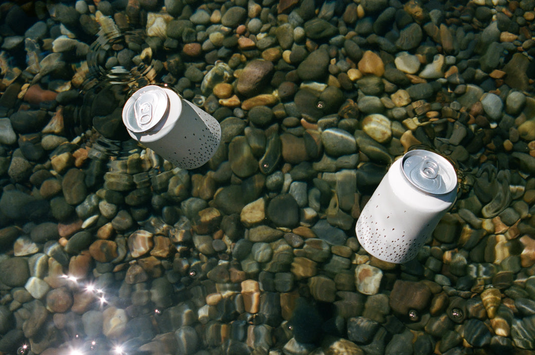 Two white Broc Cellars Love Sparkling Can Packs float on clear water over smooth, multicolored stones, with sunlight creating shimmering reflections.