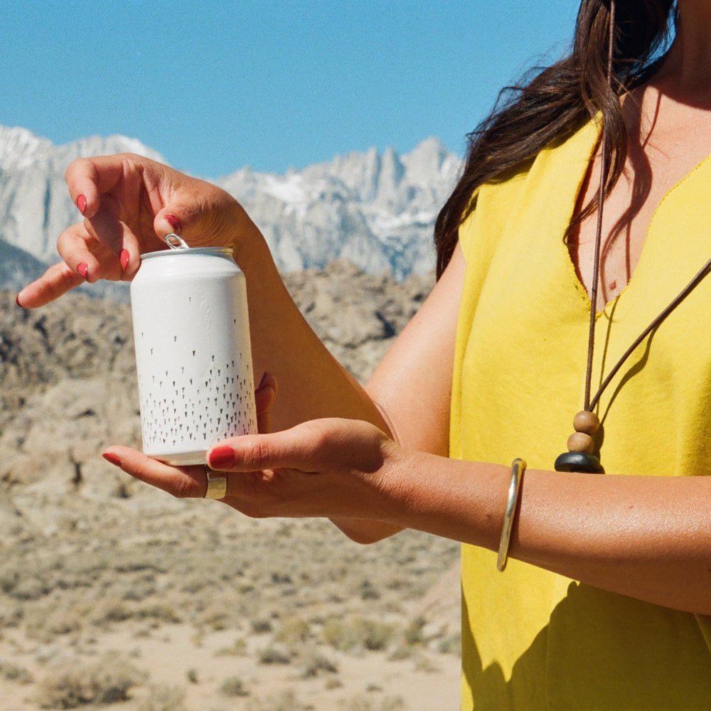 A person in a yellow sleeveless top holds a Broc Cellars Love Sparkling Can Pack—ideal for enjoying sparkling wine—amid a desert landscape with rocky terrain and snow-capped mountains in the background.