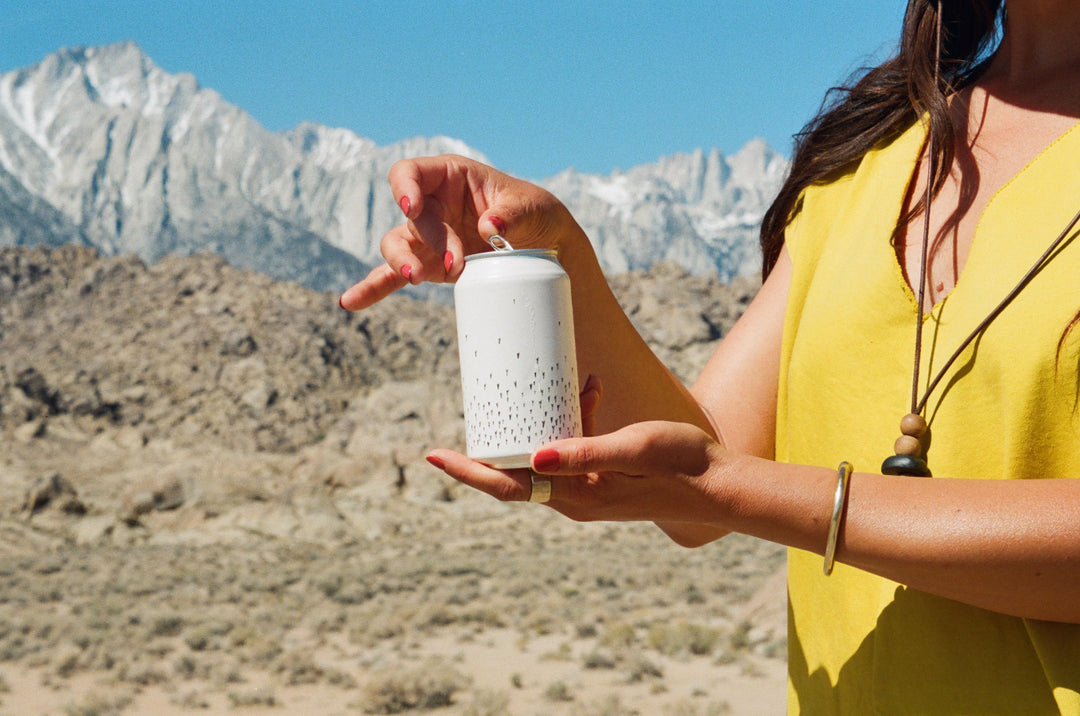 A person in a yellow sleeveless top holds a Broc Cellars Love Sparkling Can Pack—ideal for enjoying sparkling wine—amid a desert landscape with rocky terrain and snow-capped mountains in the background.