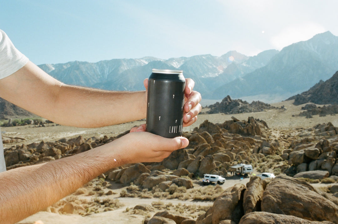 A person holds a matte black can of Broc Cellars Love Red Can Pack with both hands in a rocky desert; mountains and three parked vehicles appear in the background beneath a clear sky.