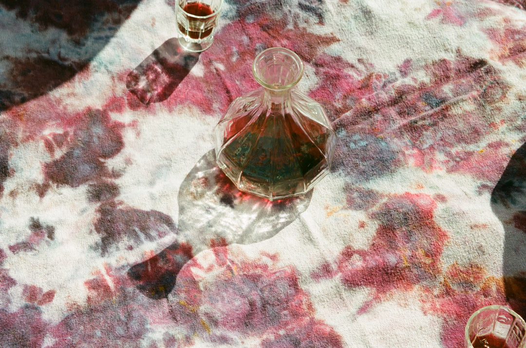 A Broc Cellars Decanter, filled with red liquid, casts a shadow on a tie-dye tablescape of pink, purple, and white hues. A small glass with the same liquid and part of another piece are seen as sunlight reflects enchantingly across the setting.