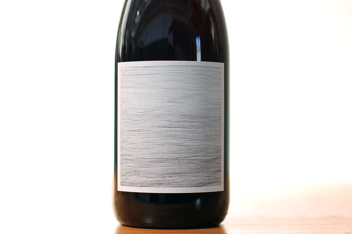 A close-up of a dark wine bottle of Broc Cellars 2018 Counoise from Mendocino, with a plain, textured white label featuring horizontal gray lines, stands on a wooden surface against a white background.