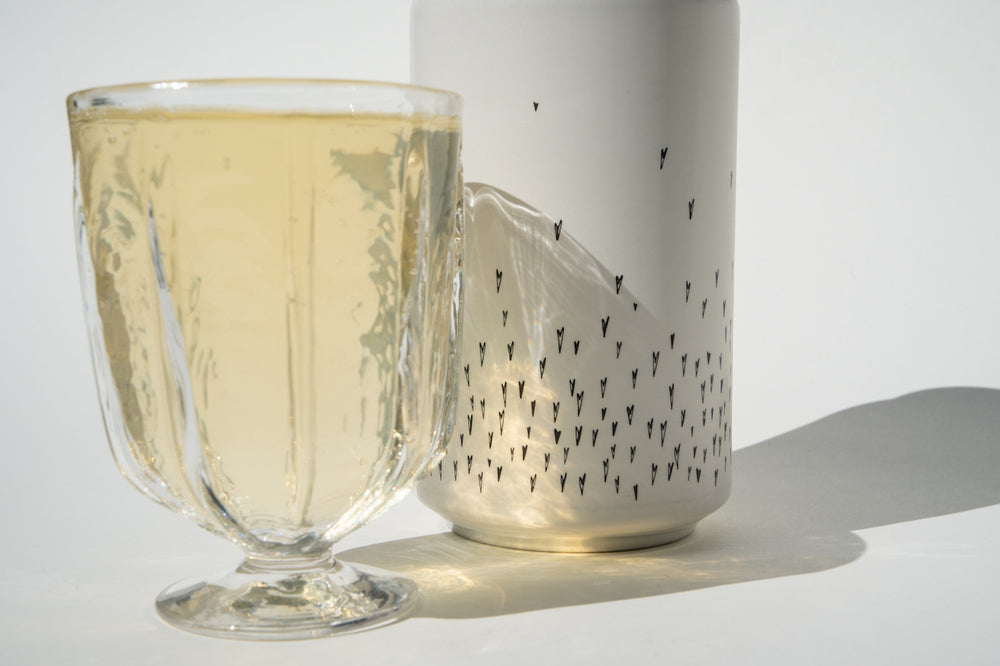 A clear glass of pale yellow Love Sparkling wine from Broc Cellars stands before a white Love Sparkling Can Pack adorned with small black fly illustrations, both casting shadows on the white surface.