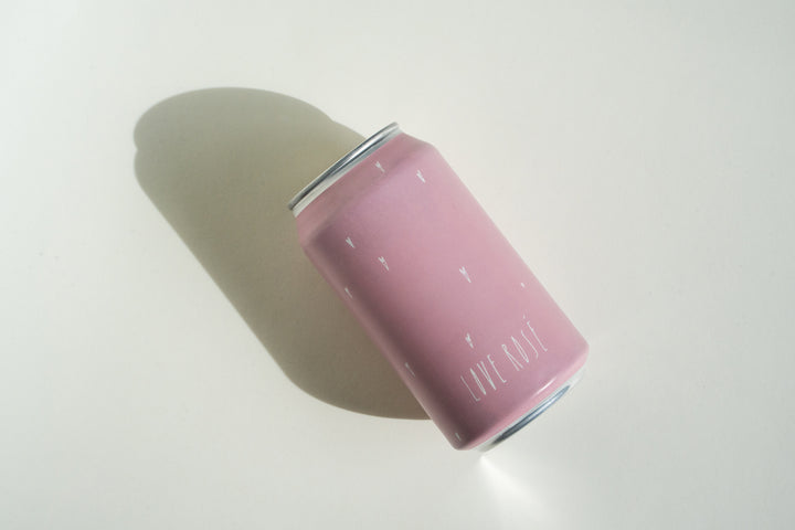 A pink aluminum Love Rosé Can by Broc Cellars, with minimal white markings and "Love Rosé" on it, lies on its side on a light surface, casting a shadow—hinting at refreshing foot-stomped Valdiguié rosé inside.