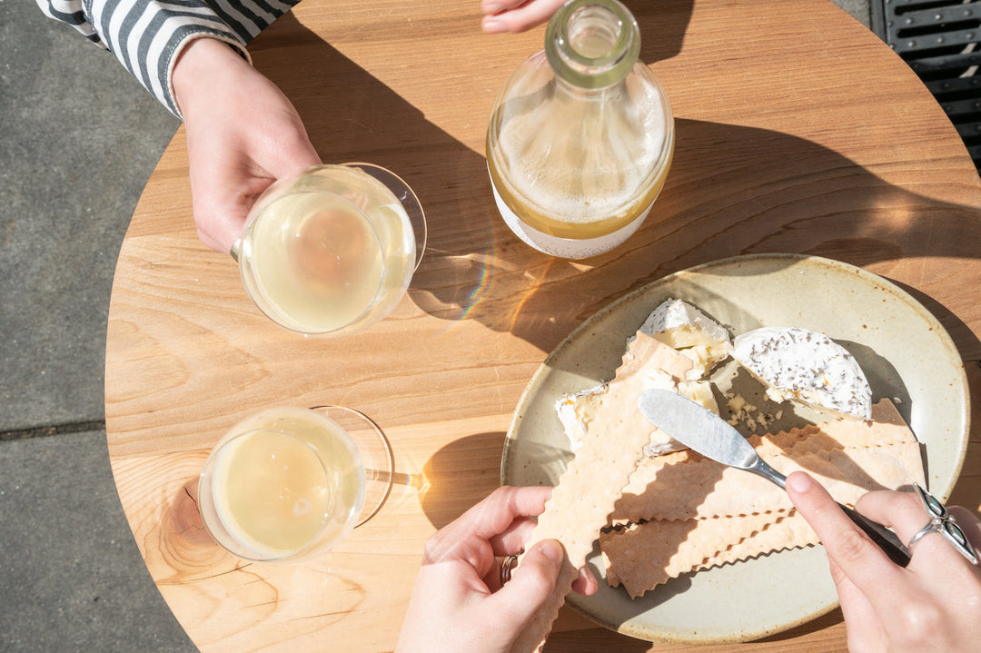 A round wooden table with Broc Cellars 2024 Love Sparkling Chenin, two glasses of sparkling white wine, an open bottle, a plate of crackers and soft cheese, and three hands—one spreading cheese, two holding glasses in natural sunlight.