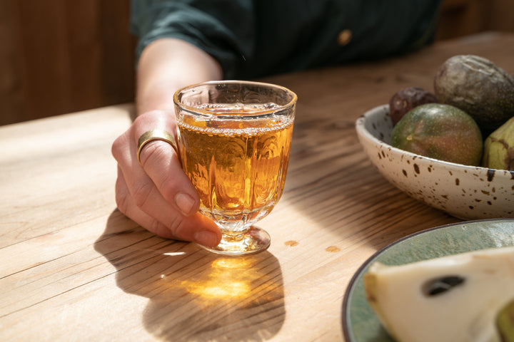 A person casually holds a Broc Glass filled with amber cider on a wooden table. Nearby, sunlight highlights the elegance of Broc Cellars handblown glassware, accompanied by a shallow ceramic bowl with assorted fruits and a plate featuring a sliced pear.