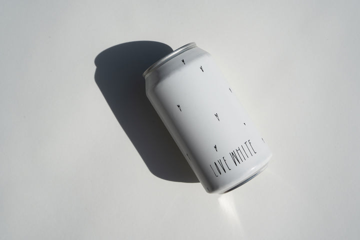 A plain white aluminum Love White Can Pack by Broc Cellars, featuring minimal black text and subtle marks on its surface, rests on a white background and casts a shadow to the left—hinting at California white wine inside.