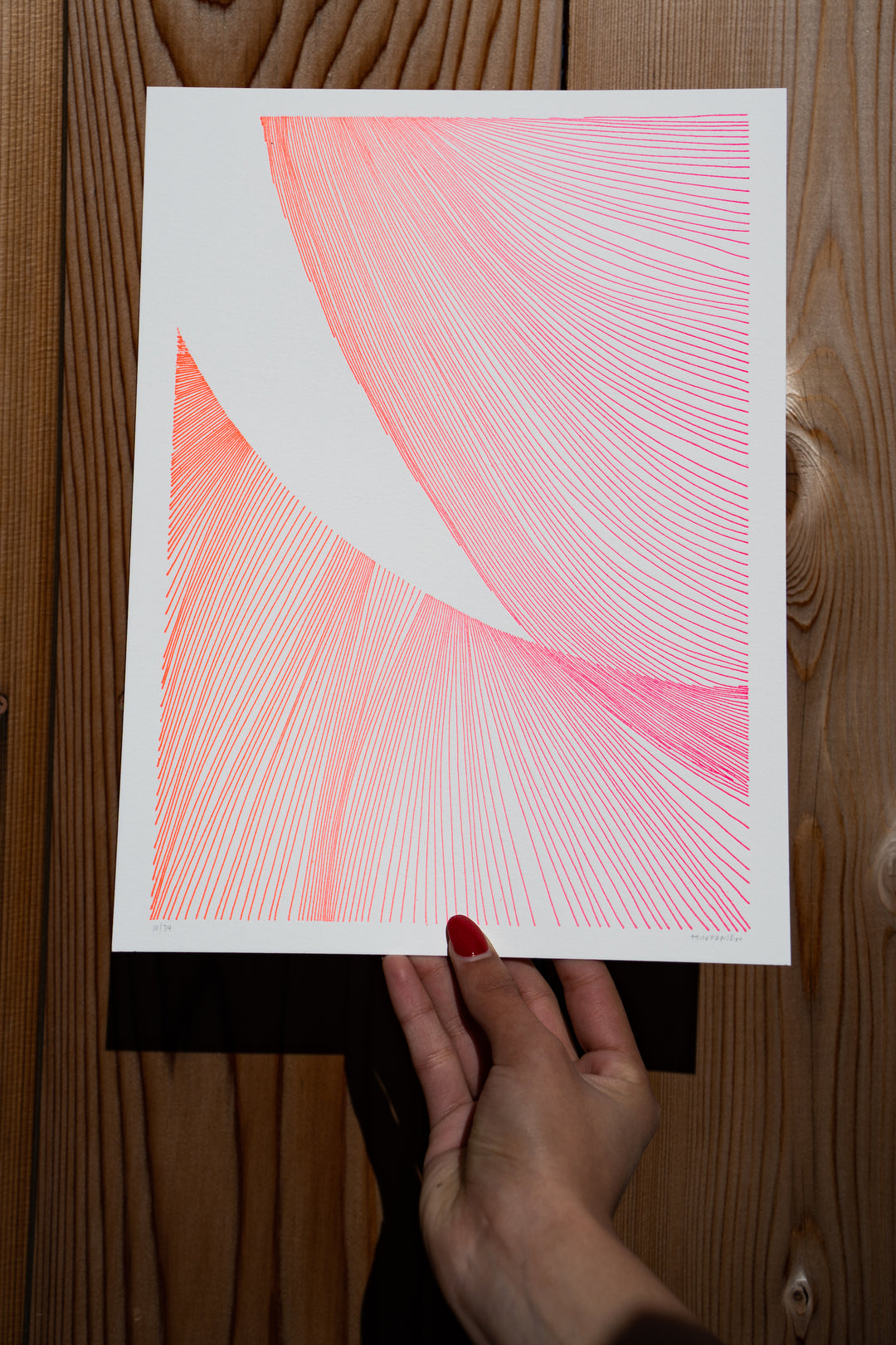 A hand with red-painted nails holds Martas Prints, a limited edition abstract pink and orange line art on premium cotton rag by Broc Cellars. The textured wooden surface in the background complements its vibrant hues perfectly.
