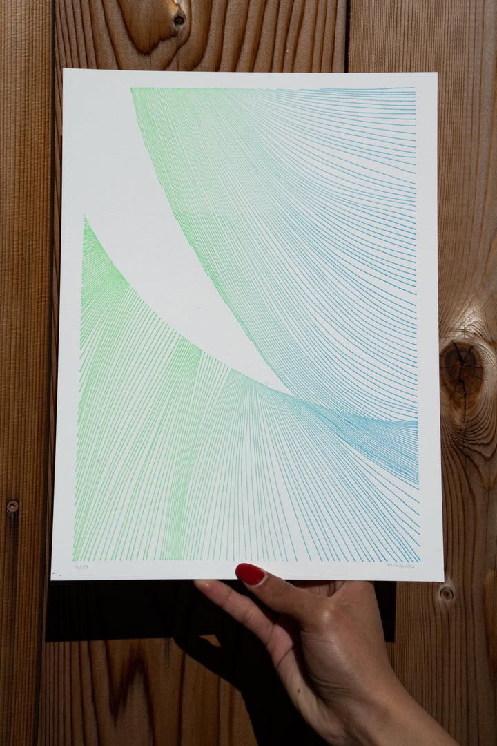 A hand with red nail polish holds a piece of paper displaying Martas Prints by Broc Cellars, featuring abstract green and blue line art with curved geometric patterns on luxurious cotton rag. The scene is set against a wooden background.