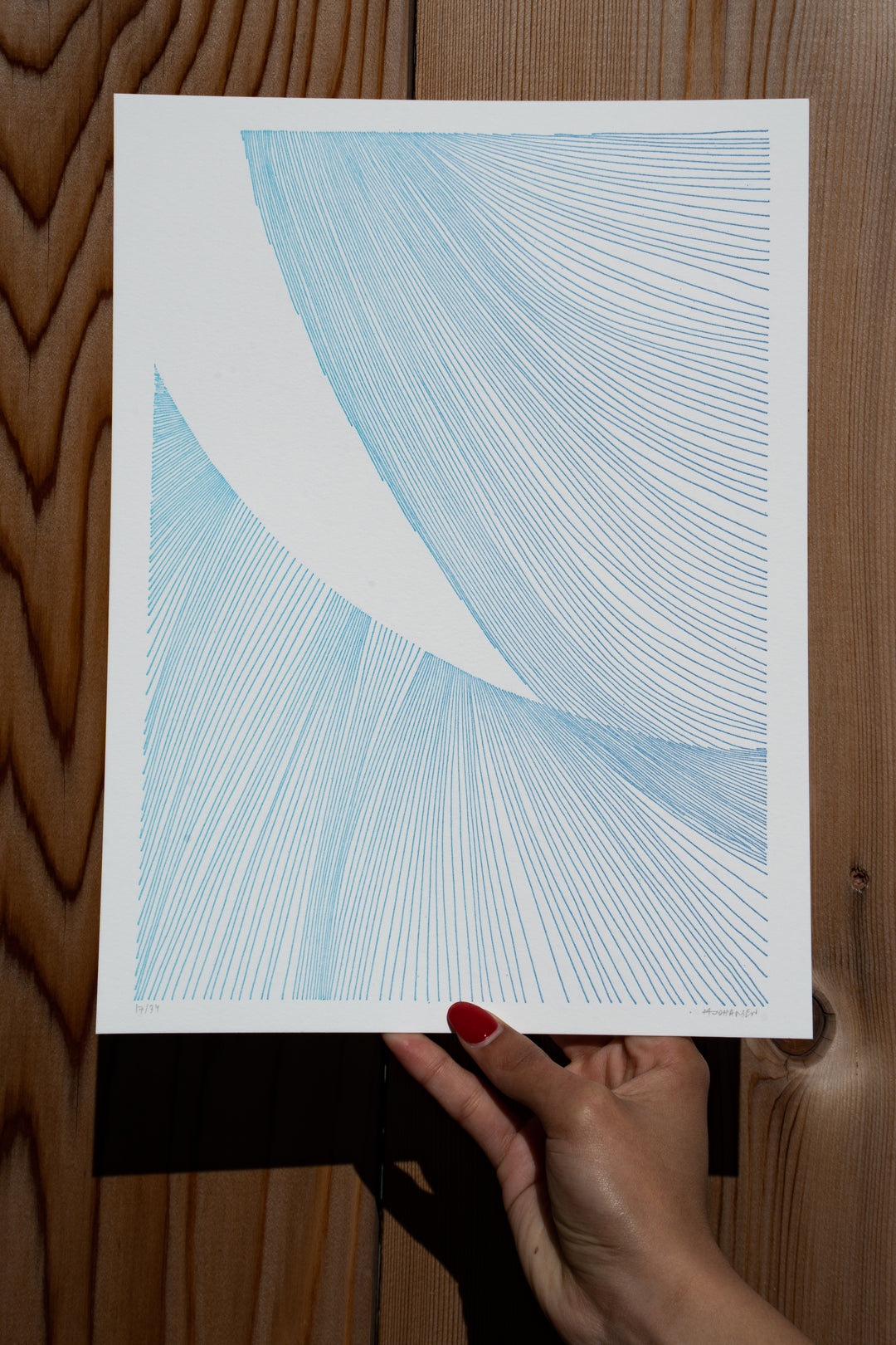 A hand with red nail polish holds a paper featuring abstract blue line art with curved patterns against a wooden background—a signature piece from Martas Prints by Broc Cellars, limited edition on premium cotton rag.