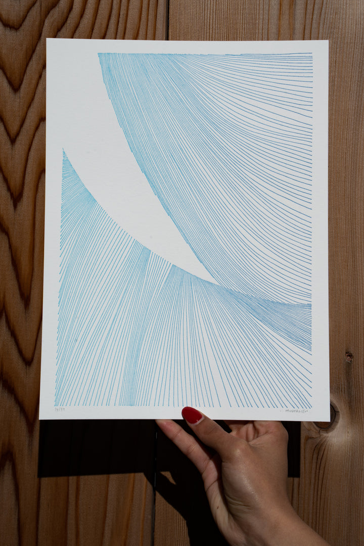A hand with red nail polish holds a paper featuring abstract blue line art with curved patterns against a wooden background—a signature piece from Martas Prints by Broc Cellars, limited edition on premium cotton rag.