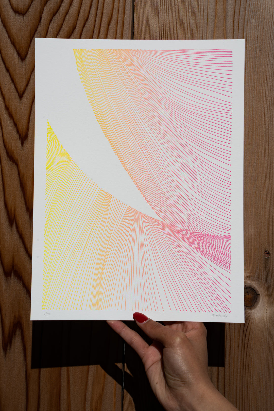 Red-polished nails hold a piece of Martas Prints from Broc Cellars against a wooden wall. Designed by Marta Johansen, it features intersecting lines in yellow, orange, and pink on luxurious cotton rag, showcasing a gradient effect. Its an exclusive part of her limited edition collection.