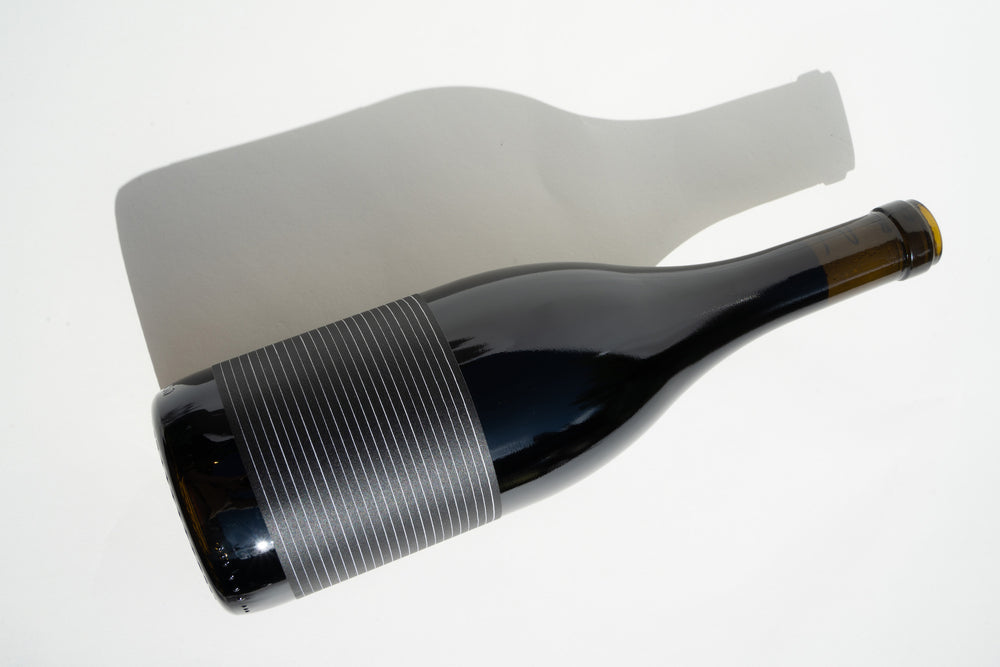 A bottle of 2019 Broc Valdiguié by Broc Cellars, featuring a minimalist striped label, rests on a white surface with its shadow cast to the side.