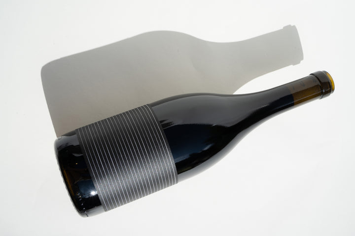 A bottle of 2019 Broc Valdiguié by Broc Cellars, featuring a minimalist striped label, rests on a white surface with its shadow cast to the side.