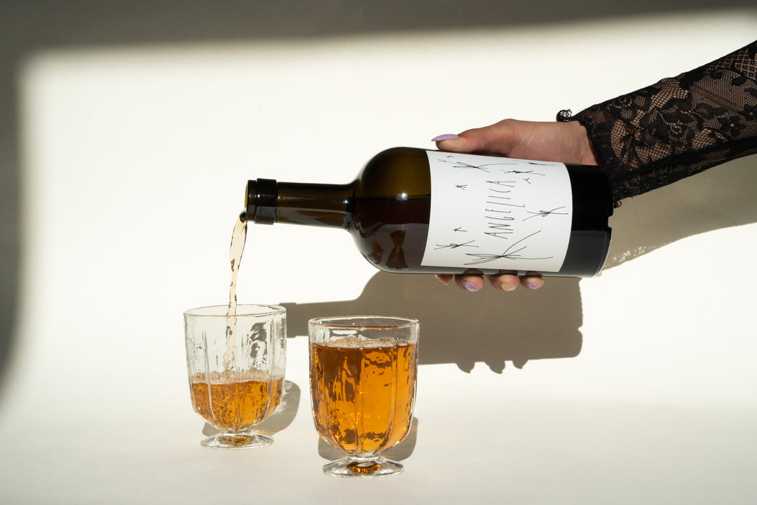 A hand with a lace sleeve elegantly pours a golden liquid from a bottle of 2020 Angelica by Broc Cellars, featuring a white label, into one of two clear glasses on a pristine white surface. A charming diagonal shadow is cast in the background.