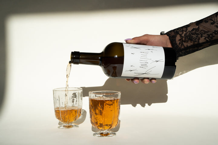 A hand with a lace sleeve elegantly pours a golden liquid from a bottle of 2020 Angelica by Broc Cellars, featuring a white label, into one of two clear glasses on a pristine white surface. A charming diagonal shadow is cast in the background.