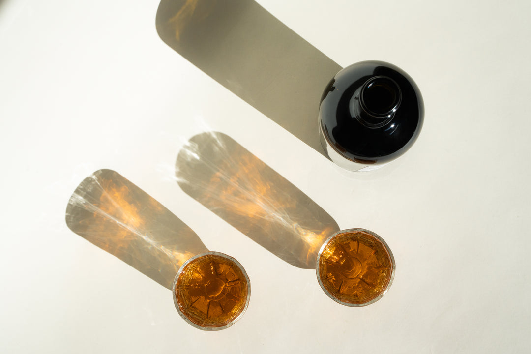 A dark bottle of Broc Cellars 2020 Angelica lies on its side on a light surface, casting a long shadow. In front, two glasses filled with this dessert wine made from Mission grapes display elongated shadows and create a sparkling effect in the light.