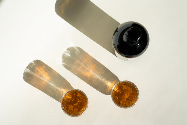 A dark bottle of Broc Cellars 2020 Angelica lies on its side on a light surface, casting a long shadow. In front, two glasses filled with this dessert wine made from Mission grapes display elongated shadows and create a sparkling effect in the light.