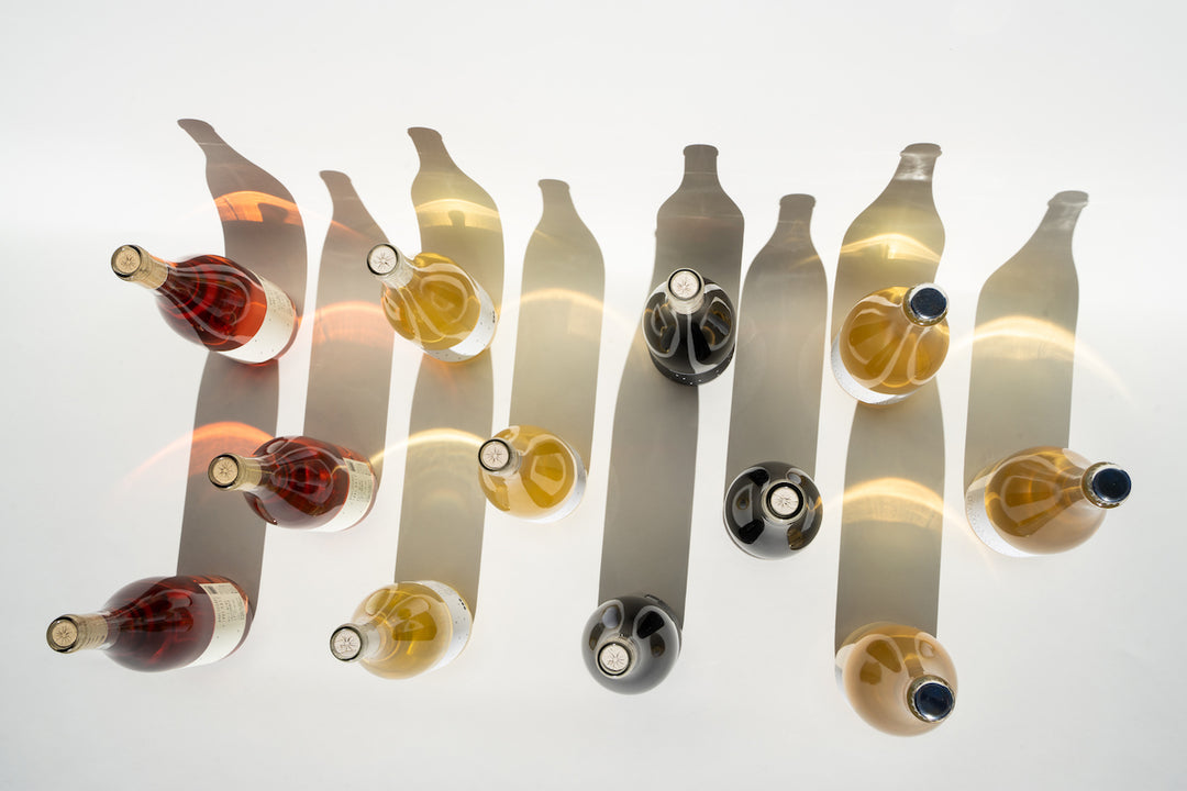 On a white surface, the Broc Cellars Love Case collection features bottles casting bottle-shaped shadows. Among them, Love Rosé and Love Sparkling shine with vibrant red, amber, and clear liquids, neatly aligned in three rows.