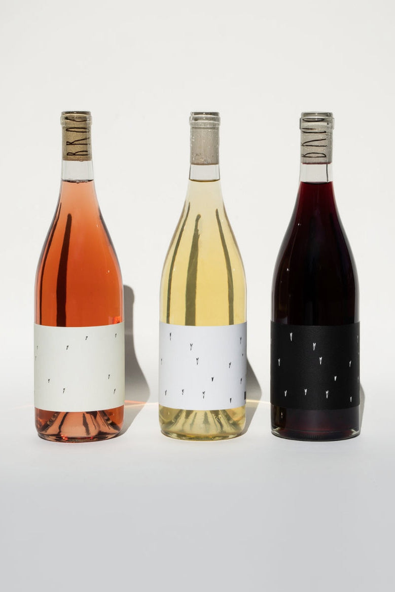 Three wine bottles from the Broc Cellars Love Trio are shown against a neutral backdrop. From left to right are: Love Rosé and Love White, both with white labels, and a red wine with a black label. Each bottle features a cork wrapped in text.