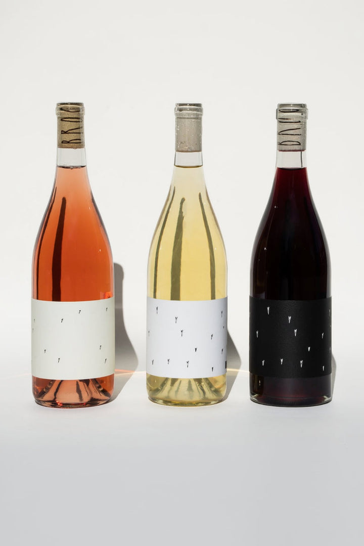 Three wine bottles from the Broc Cellars Love Trio are shown against a neutral backdrop. From left to right are: Love Rosé and Love White, both with white labels, and a red wine with a black label. Each bottle features a cork wrapped in text.