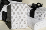 Three gifts are wrapped in Broc Cellars Conifer paper, featuring white backgrounds with black evergreen tree patterns and tied with black ribbons. The varying gift sizes highlight the unique wrapping design, ideal for gift giving.