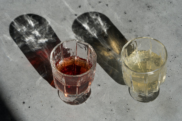 Two Broc Glass handblown glasses by Broc Cellars feature faceted textures casting patterns on a gray surface. One, similar to wine glassware, holds dark red liquid; the other has a light yellow drink. Their distinct long shadows create an effortlessly chic, casual style.