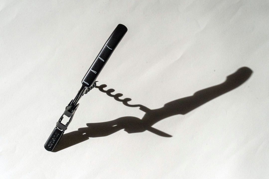 A Broc Cellars Black Wine Key with a matte black handle casts a shadow on a white surface, highlighting the spiral of the corkscrew and emphasizing the striking contrast between it and its background through dramatic lighting.