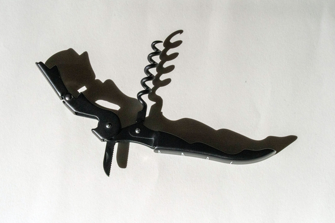 A Broc Cellars Black Wine Key with a bottle opener, small knife, and lever arm rests on a white surface, casting a shadow as its corkscrew spiral extends upward.