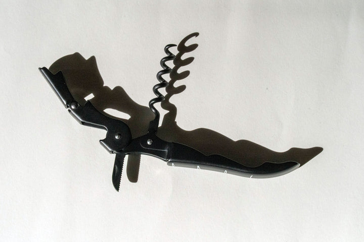 A Broc Cellars Black Wine Key with a bottle opener, small knife, and lever arm rests on a white surface, casting a shadow as its corkscrew spiral extends upward.