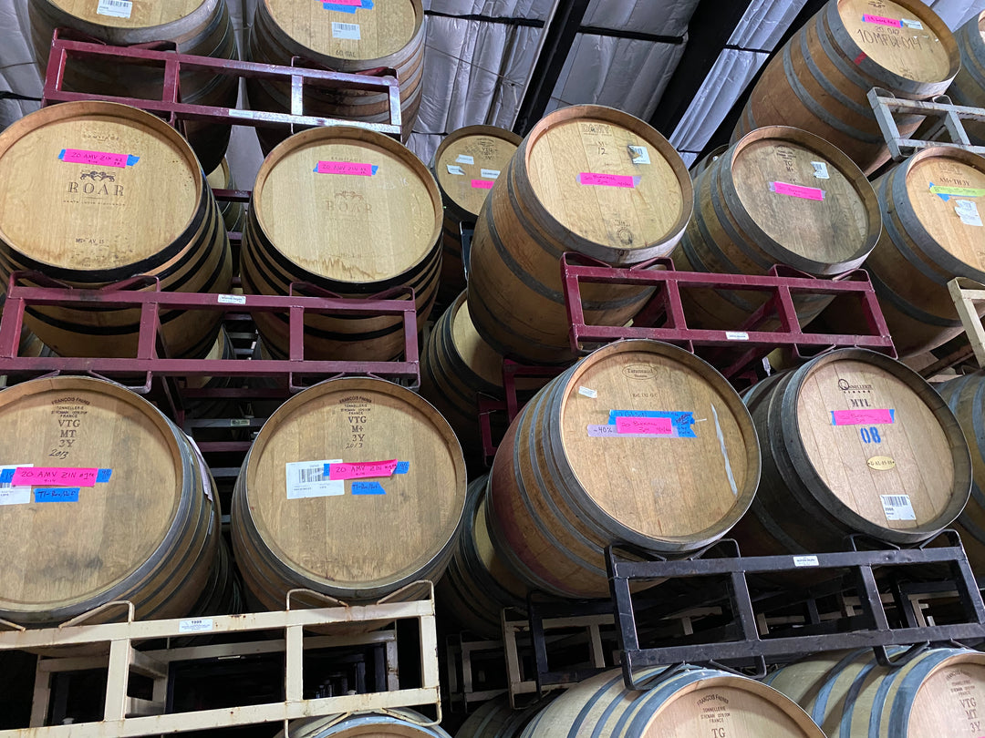 Several large wooden barrels are stacked on metal racks in a warehouse at Eaglepoint Ranch, many with pink and blue labels for Broc Cellars&