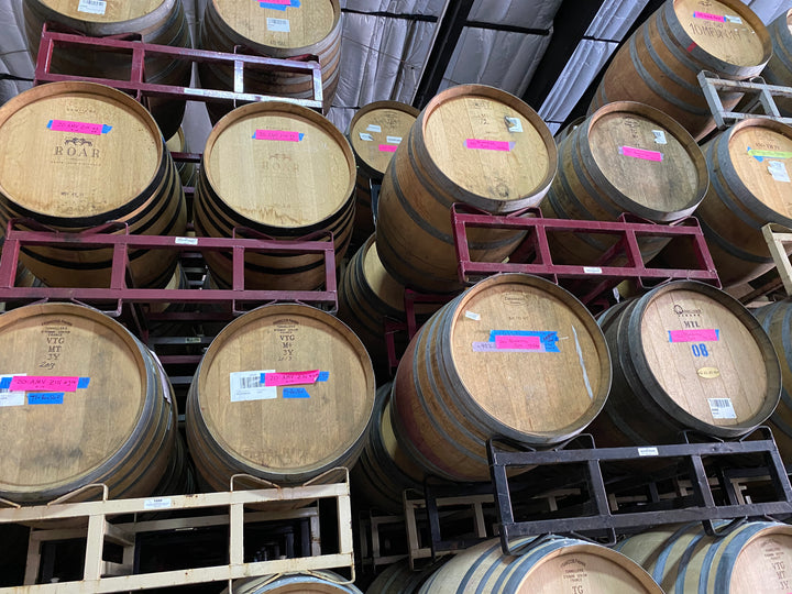 Several large wooden barrels are stacked on metal racks in a warehouse at Eaglepoint Ranch, many with pink and blue labels for Broc Cellars&