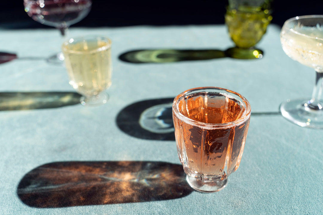 Broc Cellars Broc Glass collection features handblown glasses on a light blue surface, with a pink beverage in the foreground. Each glass casts long shadows, emphasizing the play of light on the textured surface.