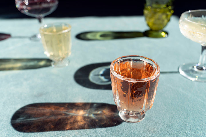 Broc Cellars Broc Glass collection features handblown glasses on a light blue surface, with a pink beverage in the foreground. Each glass casts long shadows, emphasizing the play of light on the textured surface.