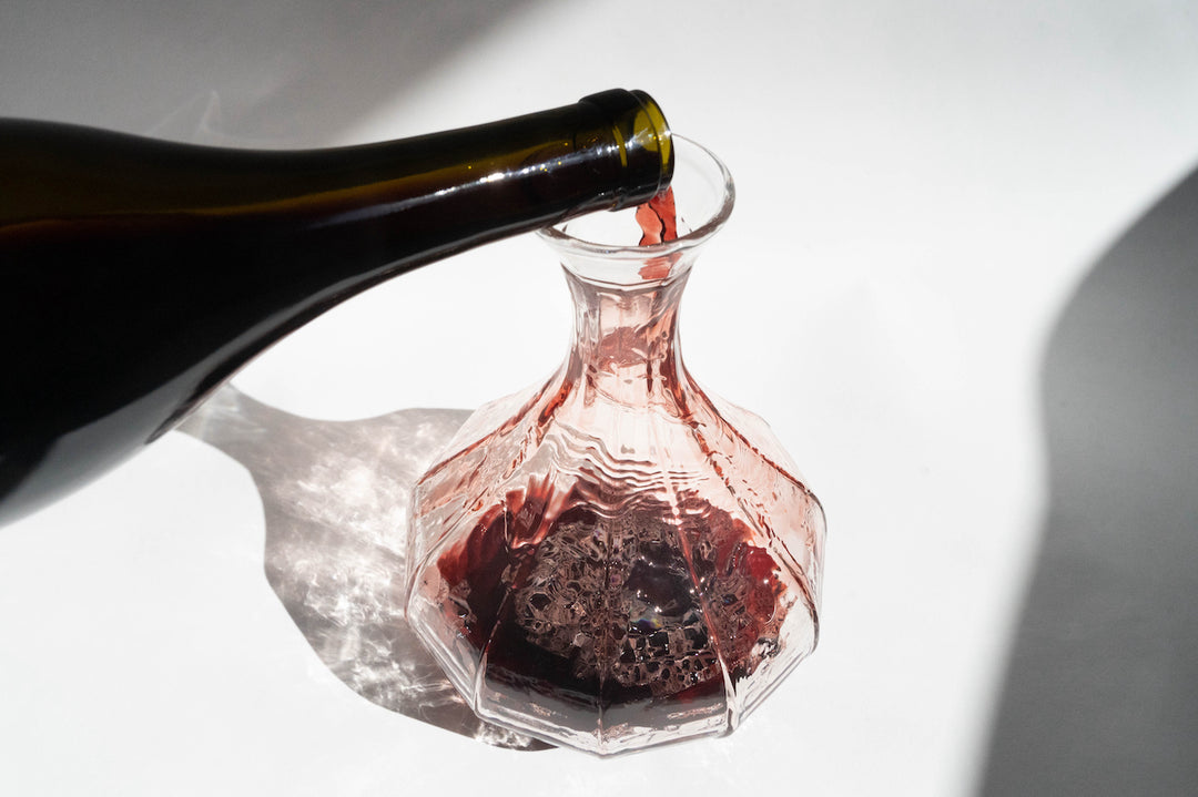 Red wine flows from a dark glass bottle into a Broc Cellars Decanter, with lighting creating strong shadows and highlighting the decanters texture, enhancing an elegant tablescape adorned with exquisite glassware.