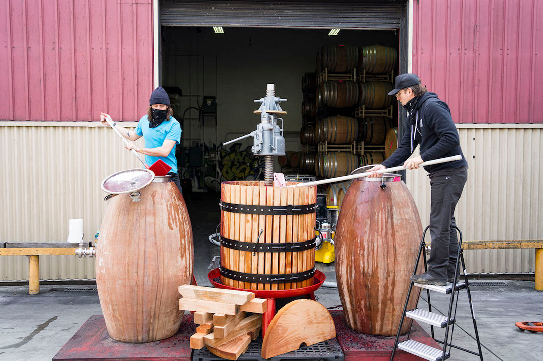 Two individuals craft wooden barrels by the warehouse entrance at Fox Hill Vineyard, home of Broc Cellars 2023 Nero D’Avola. One adjusts a lid on a stool, while another tightens metal bands on a vertical press, amidst stacked planks and barrels reflecting Mendocino craftsmanship.