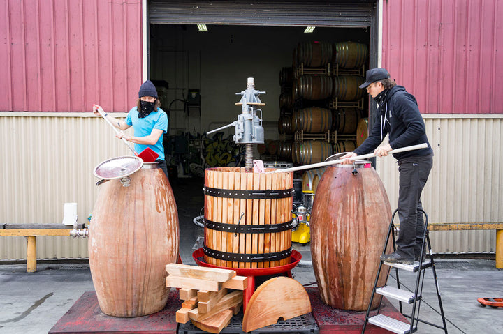 Two individuals craft wooden barrels by the warehouse entrance at Fox Hill Vineyard, home of Broc Cellars 2023 Nero D’Avola. One adjusts a lid on a stool, while another tightens metal bands on a vertical press, amidst stacked planks and barrels reflecting Mendocino craftsmanship.