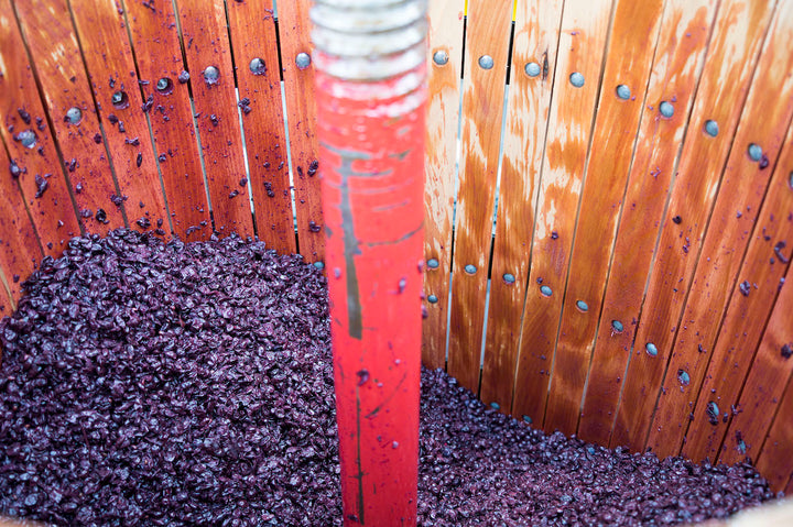 A wooden press with a central red screw mechanism is filled with crushed purple grapes from Fox Hill Vineyard for extracting juice. The wood appears stained from the grapes used to make Broc Cellars 2023 Nero DAvola wine.