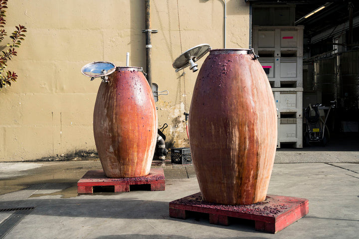 Two large wooden tanks on red pallets, potentially stained from Broc Cellars 2023 Nero dAvola production, are outdoors at Fox Hill Vineyard. Metal lids cover the cylindrical tanks, while an open warehouse door reveals stacked white containers inside.